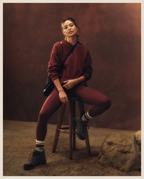 A woman poses on a wooden stool in a studio wearing a burgundy sweatshirt and leggings, black boots, and a black cross-body bag.