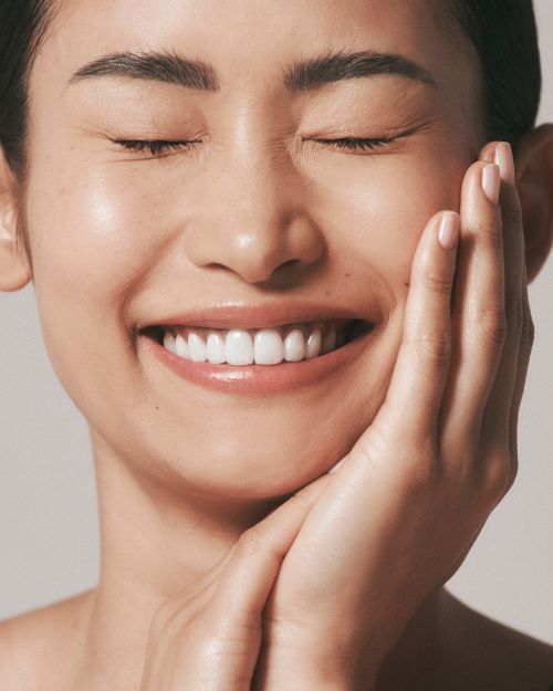 Portrait of a smiling Asian woman with closed eyes, hands on face, clear skin, and white teeth against a neutral background.