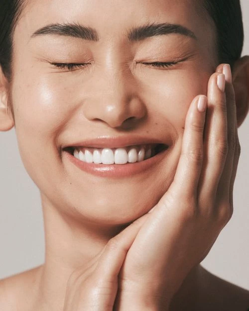 Portrait of a smiling Asian woman with closed eyes, hands on face, clear skin, and white teeth against a neutral background.