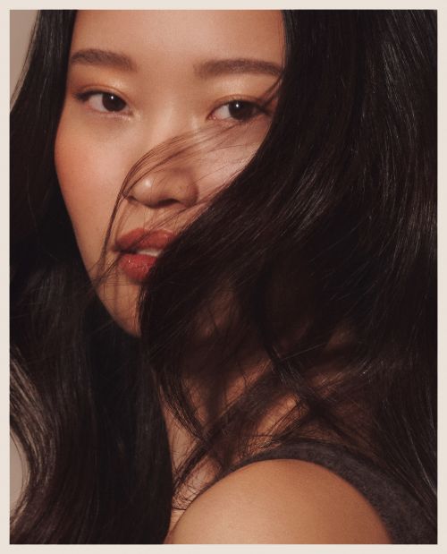 Close up portrait of an east asian woman with long dark hair blowing across her face and warm neutral makeup.