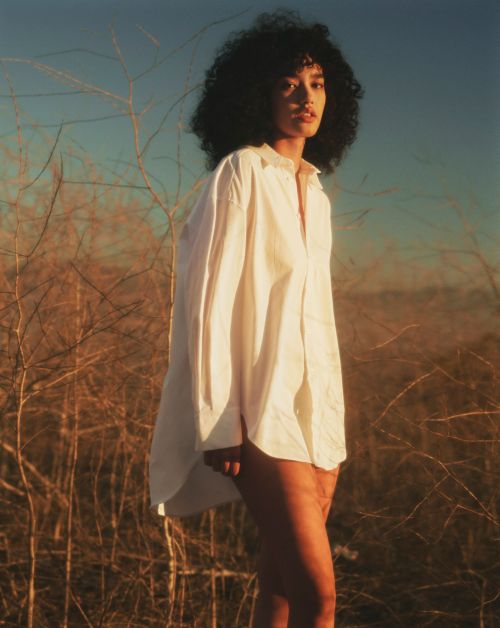Portrait of a woman with curly hair wearing a white shirt posing outdoors in a field of brown grass against a blue sky.