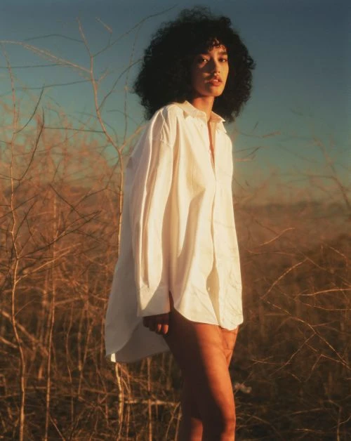 Portrait of a woman with curly hair wearing a white shirt posing outdoors in a field of brown grass against a blue sky.