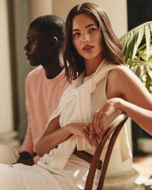 Two fashion models, an adult female and an adult male, posing outdoors in soft sunlight. The woman is seated on a chair.