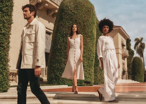 Three fashion models showcase neutral linen clothing on stone steps with lush greenery, set against a background of a building and blue sky.
