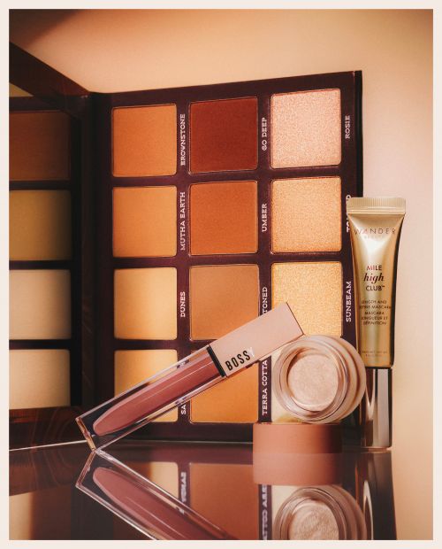 Makeup product arrangement featuring an eyeshadow palette, lip gloss, concealer, and eye cream on a reflective surface. Studio lit.