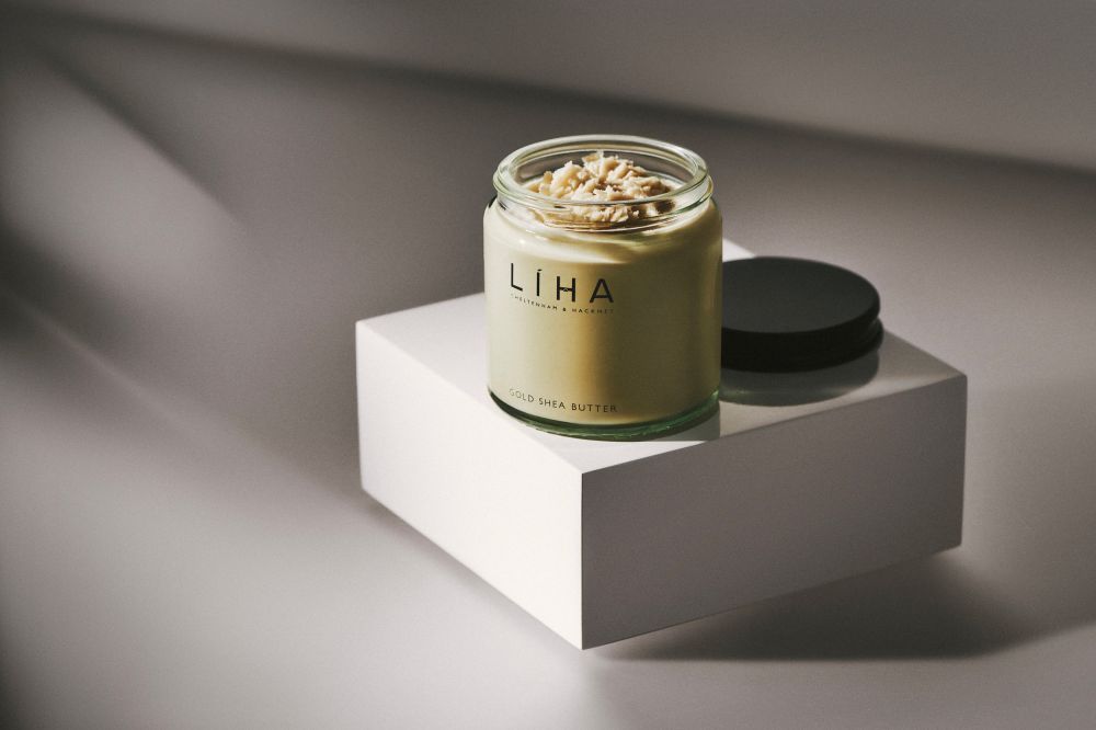 Glass jar of shea butter and lid on a white cube against a graduated gray background with soft shadows in a beauty studio setup.