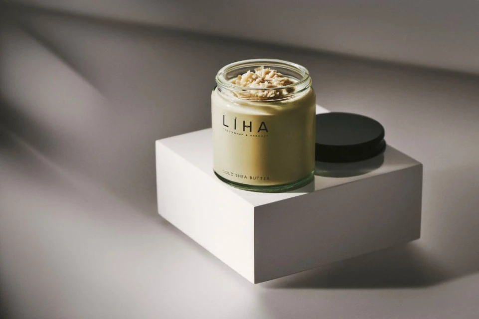 Glass jar of shea butter and lid on a white cube against a graduated gray background with soft shadows in a beauty studio setup.