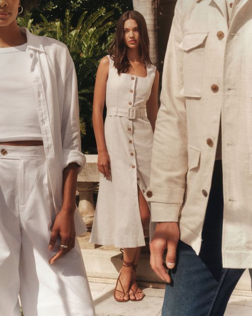 Three fashion models in casual linen outfits pose outdoors. A brunette woman wears a sleeveless dress with button-down details.