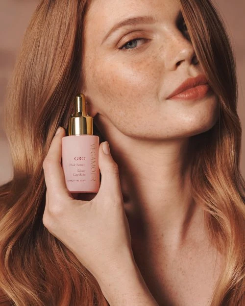 Close-up of a red-haired woman holding pink bottle of hair serum, showcasing skincare and haircare product in a beauty context.