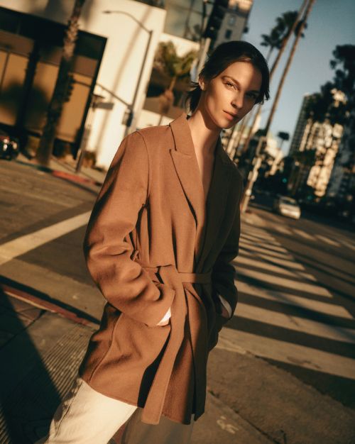 A woman in a brown blazer poses on a city street with palm trees and buildings under a bright sky, bathed in warm, directional lighting.