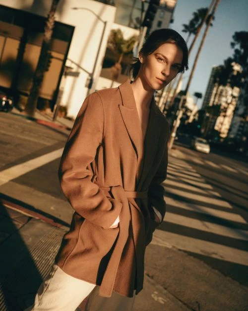 A woman in a brown blazer poses on a city street with palm trees and buildings under a bright sky, bathed in warm, directional lighting.