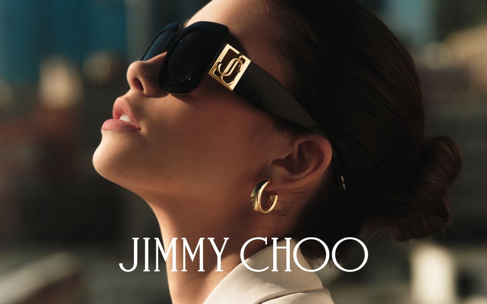 Close-up of an adult female model with dark hair pulled back, wearing black sunglasses with a gold logo, and gold hoop earrings.