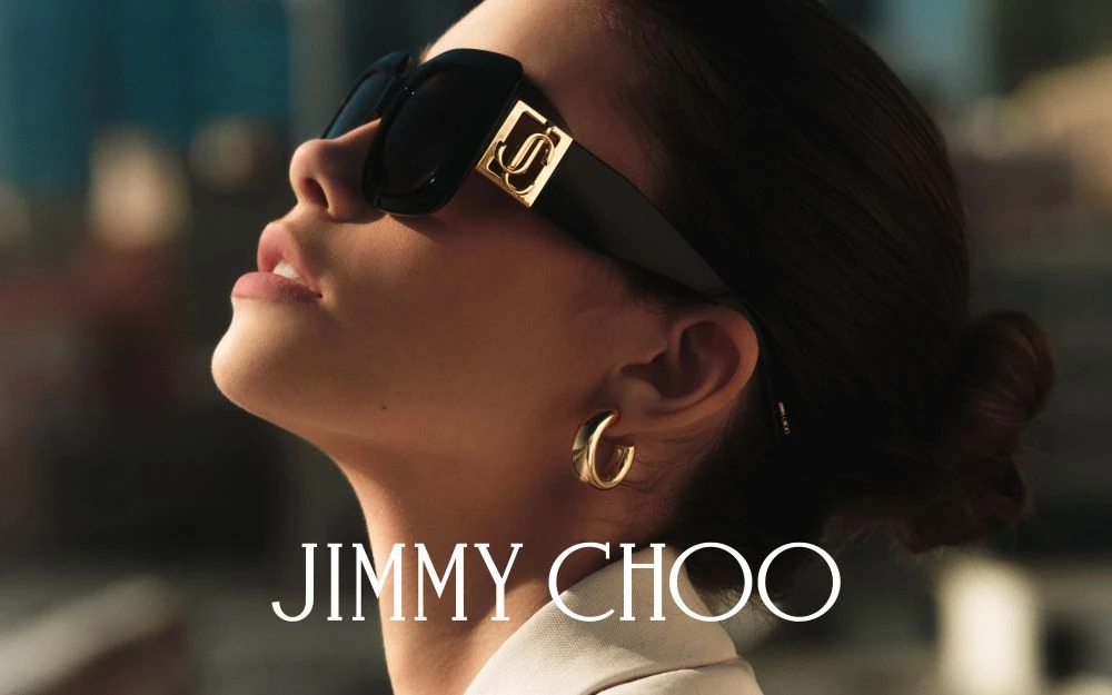 Close-up of an adult female model with dark hair pulled back, wearing black sunglasses with a gold logo, and gold hoop earrings.