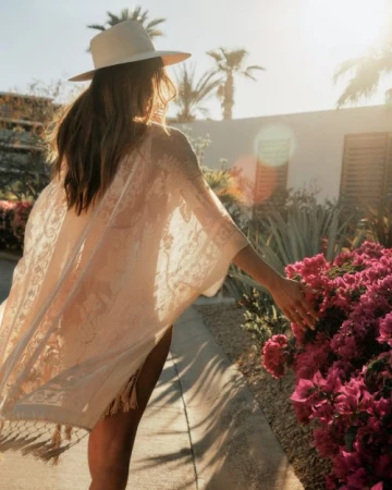 Woman with long hair wearing a lace cover-up and hat touches a flowering shrub in a garden setting on a sunny day.