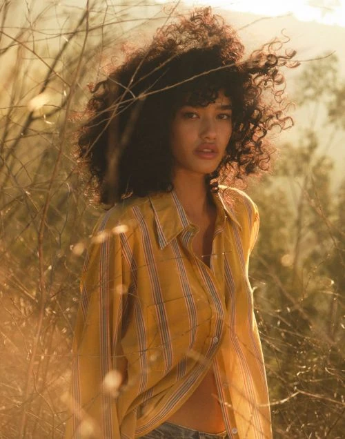 Attractive woman with curly hair posing in a yellow striped shirt outdoors among dry plants in warm, soft light creating a natural, serene mood.