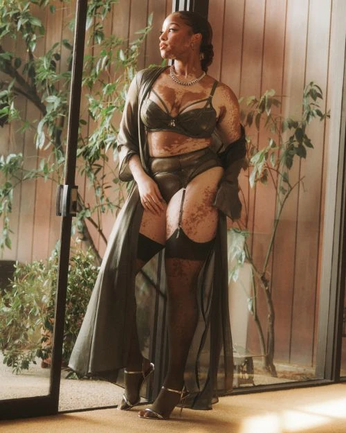 Adult female poses indoors wearing lingerie and robe. She wears open-toed heels and jewelry, framed by indoor plants and natural light.
