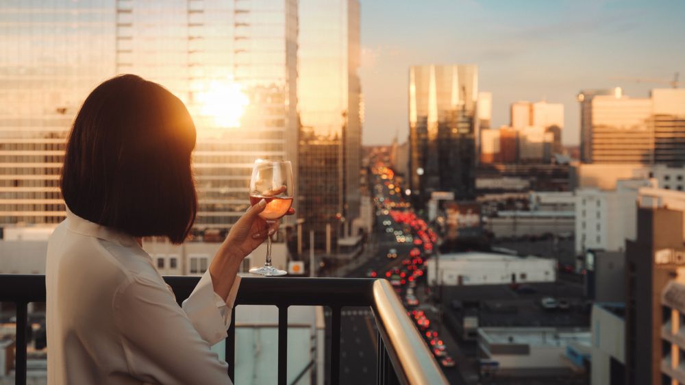 Adult female drinking wine on an apartment balcony with a city skyline view at sunset, wearing a white blouse.