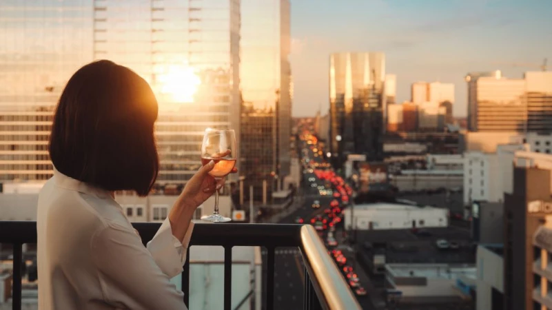 Adult female drinking wine on an apartment balcony with a city skyline view at sunset, wearing a white blouse.