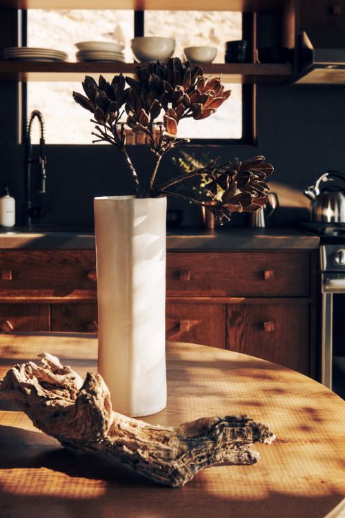 Interior kitchen scene featuring a white vase holding dried foliage, resting on a wood table, with a piece of driftwood beside it.