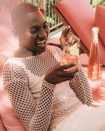 Black man with short hair wearing a cream mesh top laughing while holding a wine glass with a drink