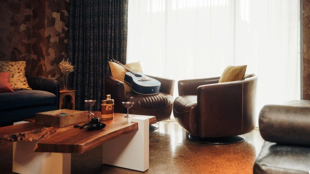 Living room interior with leather armchairs, wooden coffee table, blue sofa, guitar, and decorative objects in warm lighting.