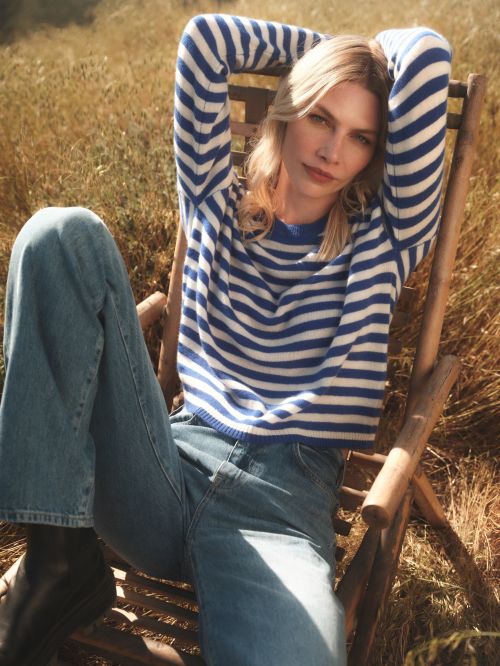 Woman in blue-and-white striped sweater and blue jeans sitting in wooden chair in field of brown grasses, posing for fashion shot.