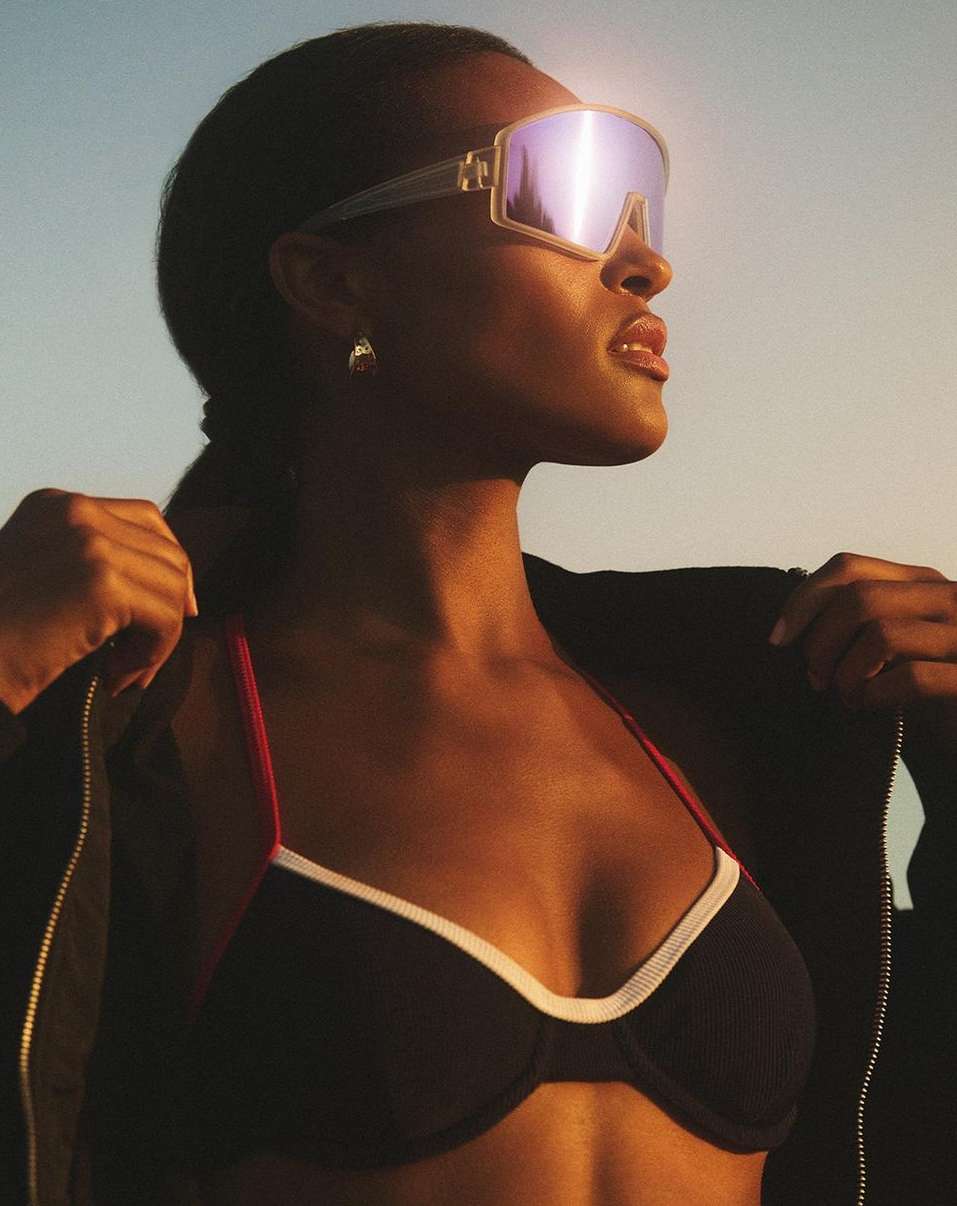 Woman in sunglasses and a sports bra poses against a blue and orange sky, wearing a jacket and gold earrings.