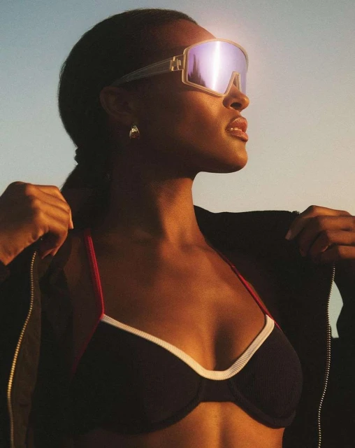 Woman in sunglasses and a sports bra poses against a blue and orange sky, wearing a jacket and gold earrings.