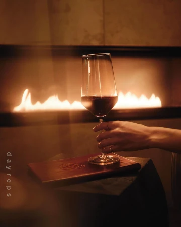 A hand holding a wine glass filled with red wine in front of a warm fireplace by Wolfe + Von.
