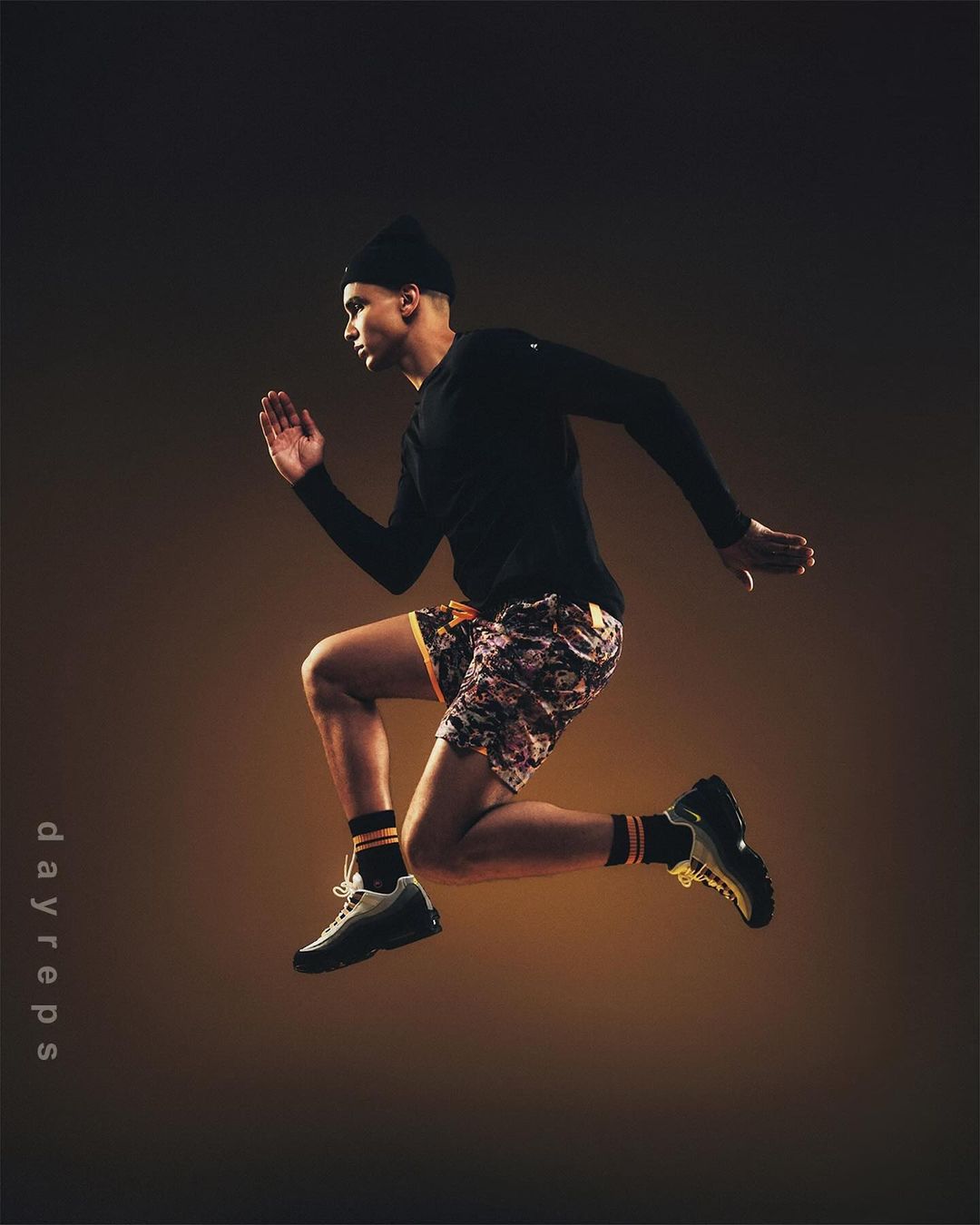 Athletic man with dark hair wearing black hat and long-sleeve shirt jumps in mid-air against a warm, brown gradient background.