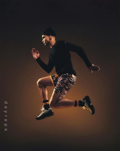 Athletic man with dark hair wearing black hat and long-sleeve shirt jumps in mid-air against a warm, brown gradient background.