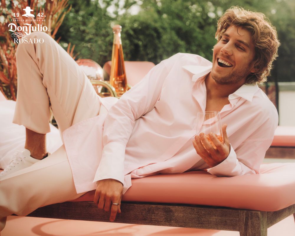 Man in pink shirt and pants lounges on a pink chair with a drink, showcasing a Don Julio Rosado tequila bottle and logo in a backyard setting.