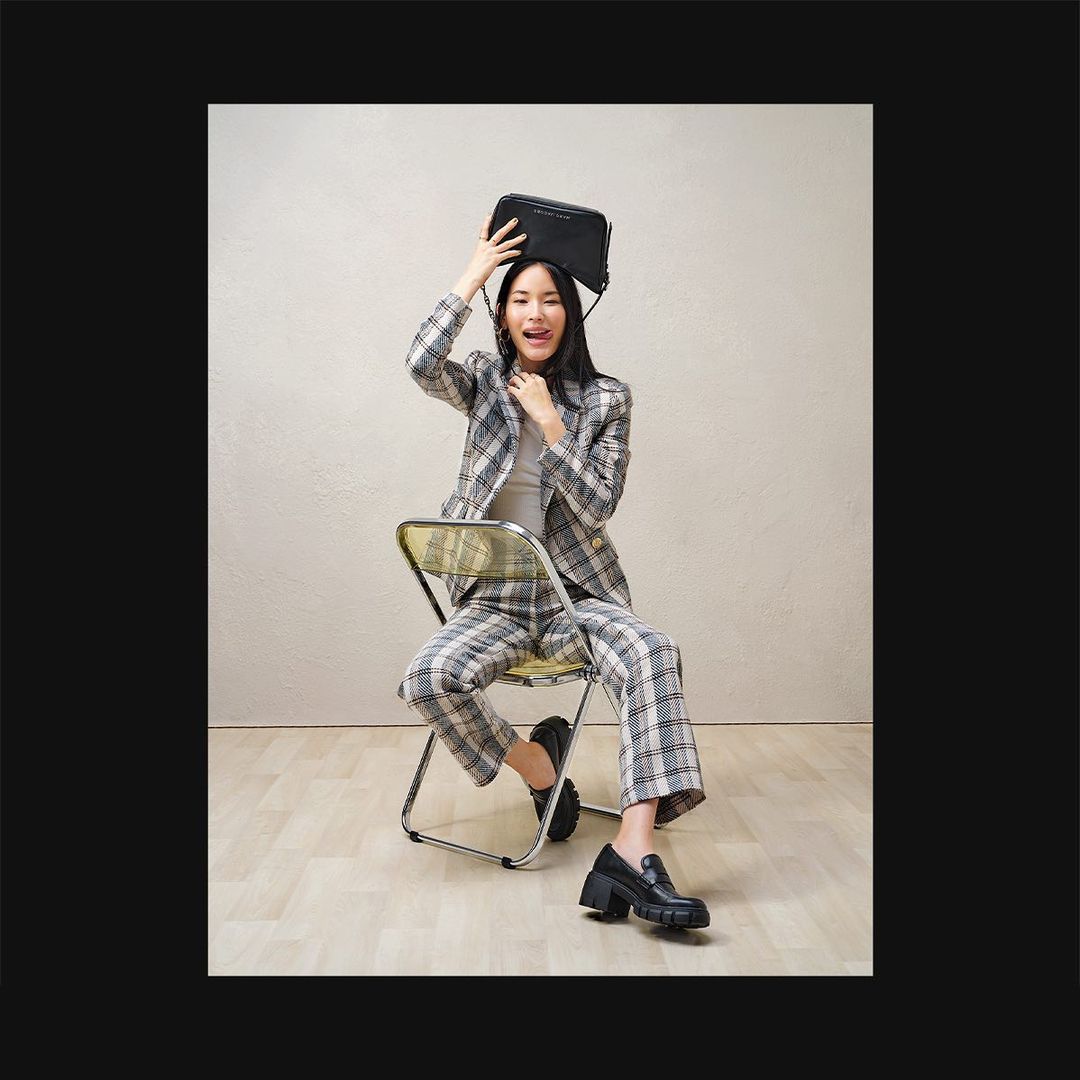 Stylish woman in plaid suit playfully poses with black bag on head, seated indoors with neutral backdrop, showcasing fashion.