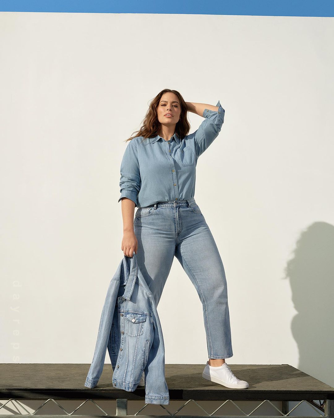 Ashley Graham poses in a denim shirt and jeans, holding a denim jacket, in front of a white and blue background.