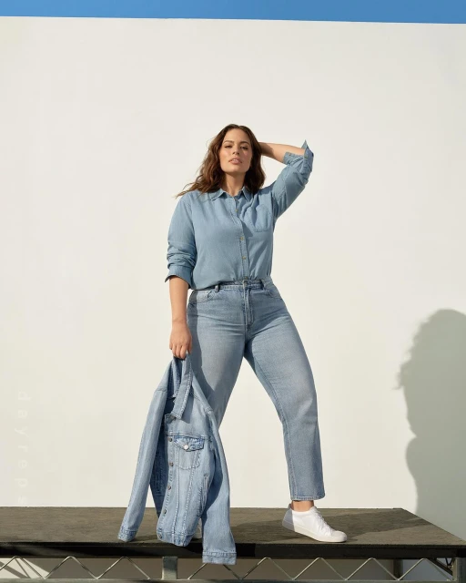 Ashley Graham poses in a denim shirt and jeans, holding a denim jacket, in front of a white and blue background.