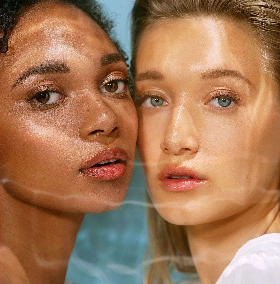 Close-up of two women with light makeup and glowing skin, posing together in front of a soft blue background with light reflecting on their faces.