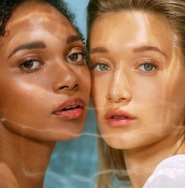 Close-up of two women with light makeup and glowing skin, posing together in front of a soft blue background with light reflecting on their faces.