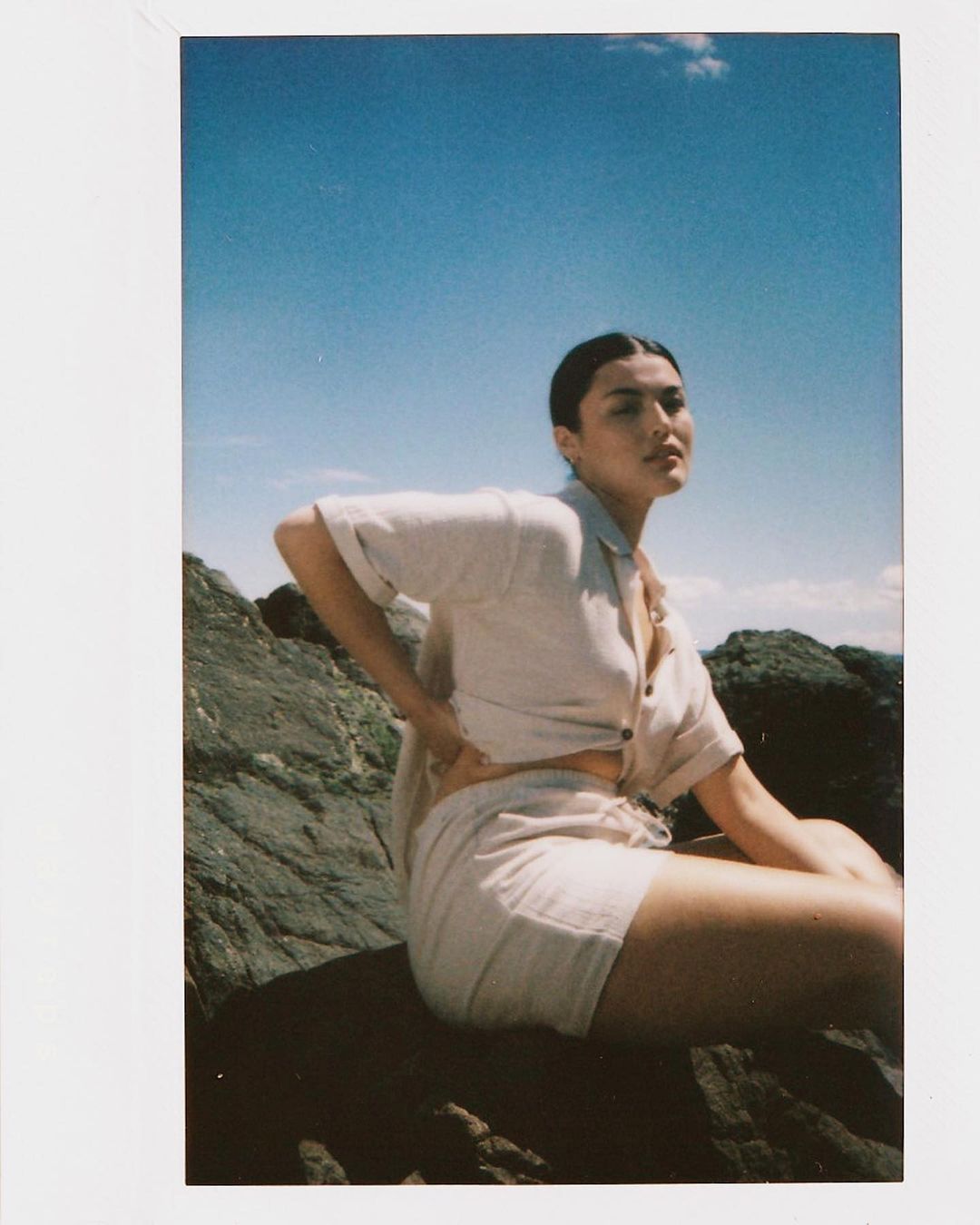 Woman in beige linen shirt and shorts sitting on a rock formation against a blue sky and ocean backdrop in a sunlit outdoor scene.