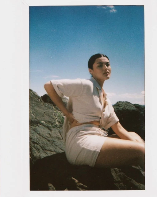 Woman in beige linen shirt and shorts sitting on a rock formation against a blue sky and ocean backdrop in a sunlit outdoor scene.
