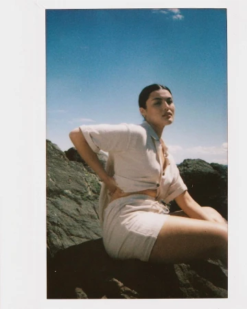 Woman in beige linen shirt and shorts sitting on a rock formation against a blue sky and ocean backdrop in a sunlit outdoor scene.