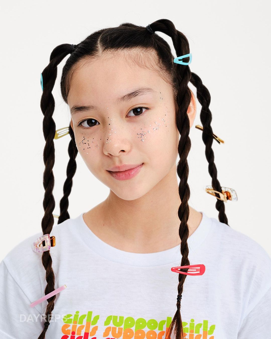 Portrait of a young Asian girl with sparkly makeup and braided hair adorned with colorful hair clips against a bright white backdrop.