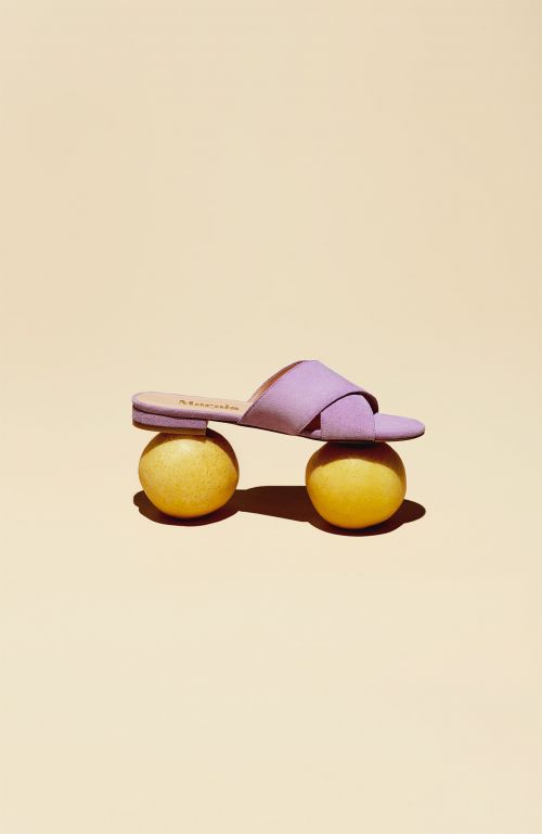 Studio shot of a single purple sandal balanced atop two yellow spherical objects against a peach-colored backdrop, product photography.