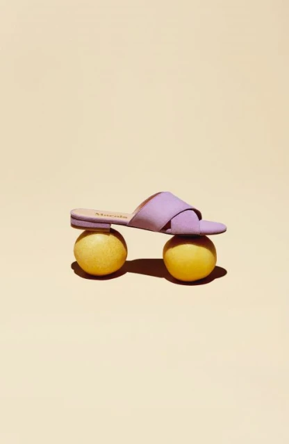 Studio shot of a single purple sandal balanced atop two yellow spherical objects against a peach-colored backdrop, product photography.