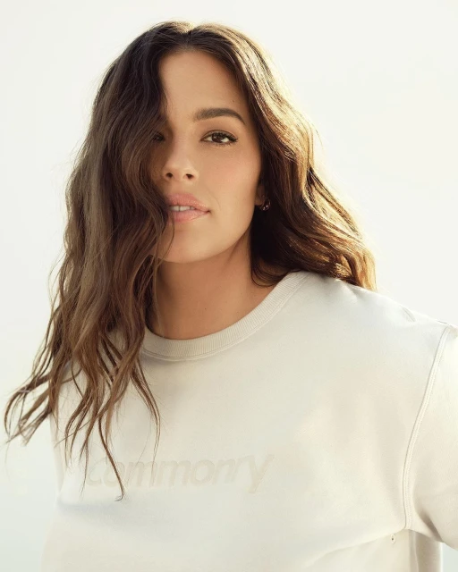 Portrait of a young woman with long wavy brunette hair wearing a light-yellow sweatshirt, posing indoors in front of a bright background.