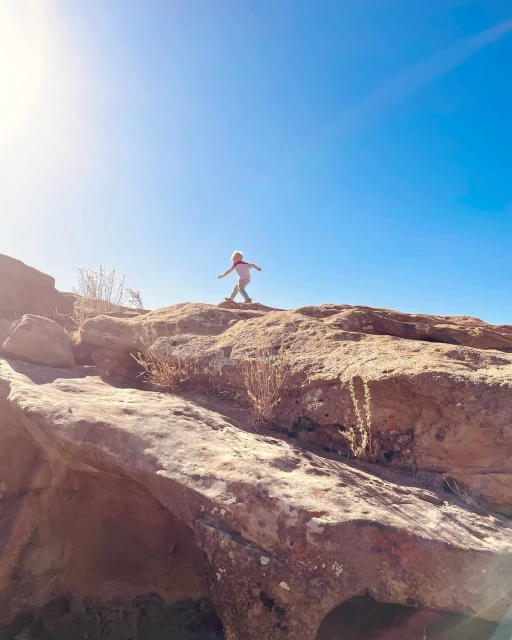 Low-angle shot of child with outstretched arms standing on rocky terrain under clear blue sky, captured during daylight.