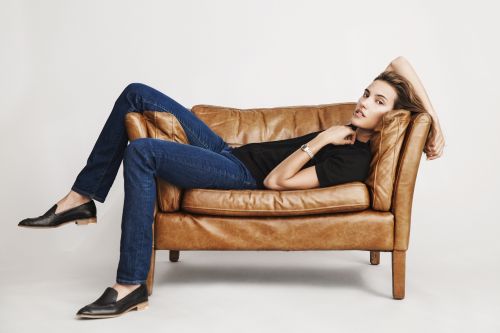 A woman lounges in a brown leather armchair, wearing jeans, a black shirt, and loafers against a light background in a studio setting.