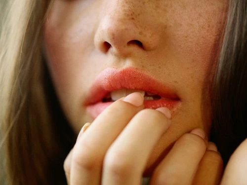 Extreme close-up portrait of a woman biting her fingernail, highlighting freckles and coral lips by Kathryna Hancock