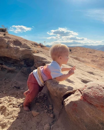 A toddler standing on bare dirt and rocks outdoors, reaching up onto a ledge under a blue sky with scattered white clouds.
