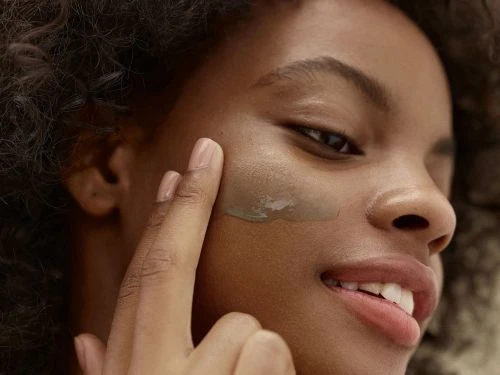 Smiling young woman applies green skincare mask with fingers for beauty treatment in a close-up beauty shot.