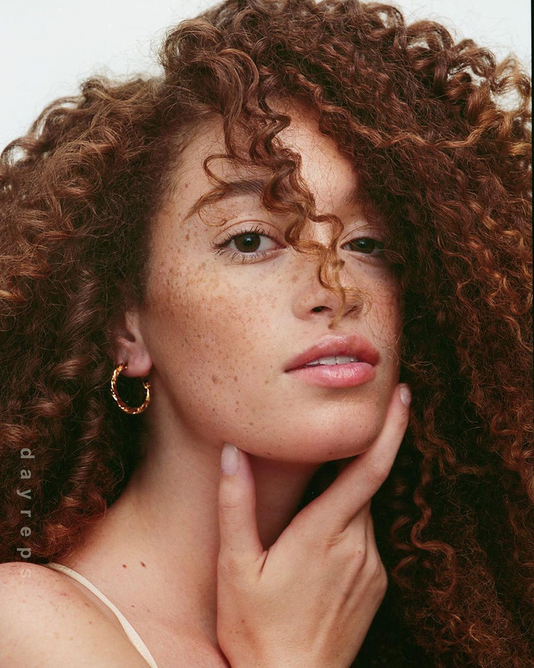 Close-up portrait of a curly-haired woman with freckles touching her face. She has light skin and is wearing gold hoop earrings.